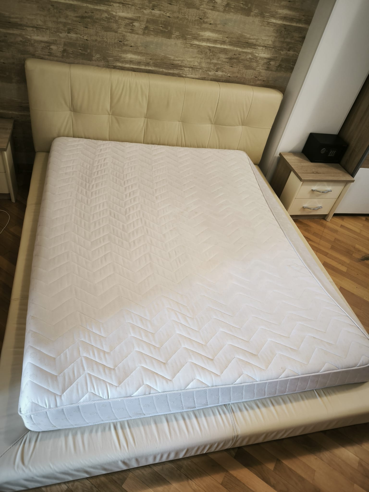 White mattress with chevron pattern on a beige leather bed frame in a wooden floored bedroom next to a nightstand with a black safe.