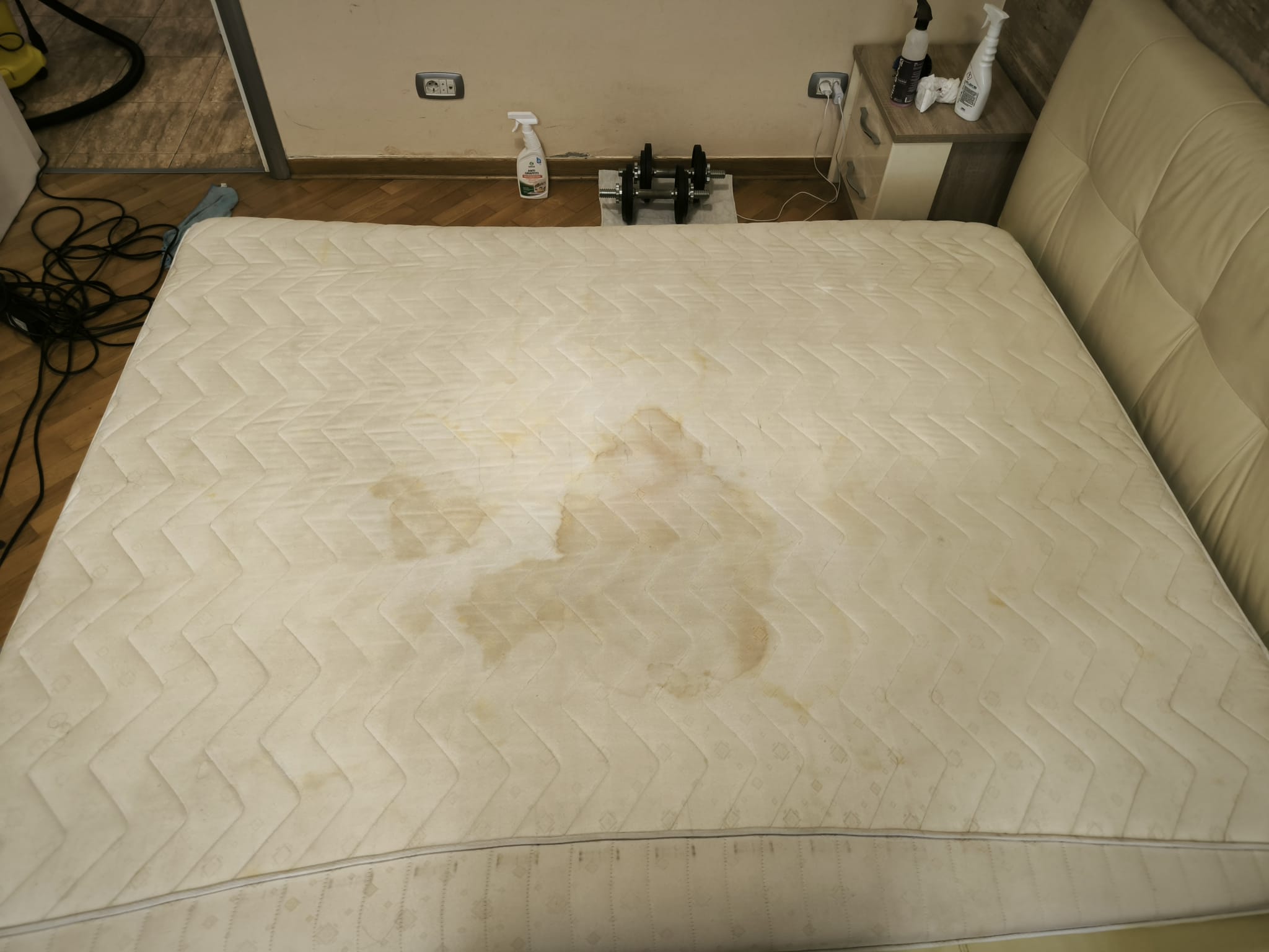 White mattress with multiple large yellowish stains, placed on a beige bed frame in a room.